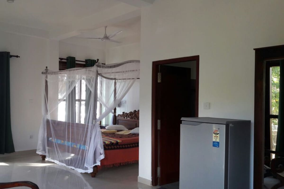 Een slaapkamer met een hemelbed, een plafondventilator, een kleine koelkast en een deur die naar een andere kamer leidt.