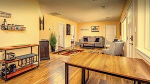 A living room with a wooden coffee table, a sofa, a grey armchair, a kitchen area with a stove, and a dining table with a metal shelf. - Newly remodeled quaint Cottage (Three Rivers)