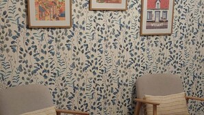 Una habitación con dos sillas de madera, una mesita de centro de vidrio, papel tapiz estampado y cuadros con vistas de la ciudad en la pared.
