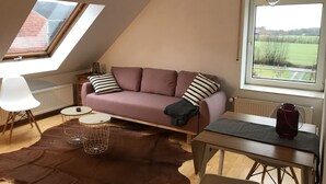 A cozy loft bedroom with a sloped ceiling, a purple sofa, a dining table, a large window with a view of a green field, and a skylight. - Apartment No. 2 in the Main House – bright, cozy, and well equipped (Havixbeck)