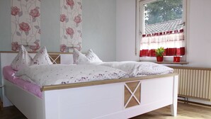 Un lit bien fait, recouvert d’une couvre-lit à motifs floraux et agrémenté d’oreillers, un tapis rouge et une fenêtre ornée d’un rideau rouge et d’une plante en pot.