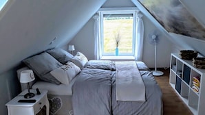 Un dormitorio con techo inclinado, una cama con ropa de cama gris, una mesita de noche con lámpara, un ventilador de pie y una ventana con vistas a zonas verdes.
