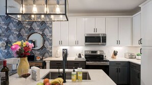 Une cuisine moderne avec des meubles blancs, un four à micro-ondes en acier inoxydable et un grand îlot central agrémenté de fruits et d’une bouteille de vin.
