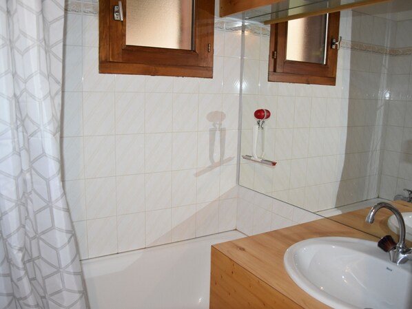 Une salle de bain avec des murs recouverts de carreaux blancs, un meuble-lavabo en bois, un lavabo, un miroir et un rideau de douche.