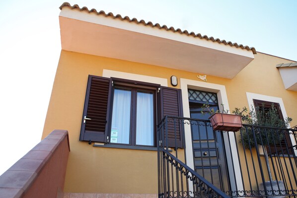 A building with a yellow facade, a tiled roof, a balcony with a metal railing, and a window with shutters.
