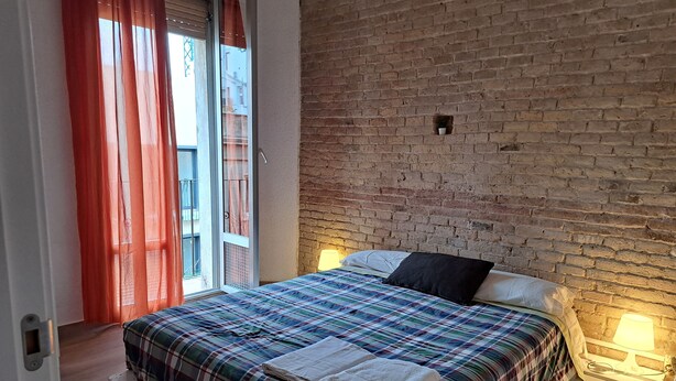 A bedroom with a brick wall, a plaid bedspread, two bedside lamps, and a window with red curtains. - Casa Mia Barcelona & Barça (Hospitalet de Llobregat)