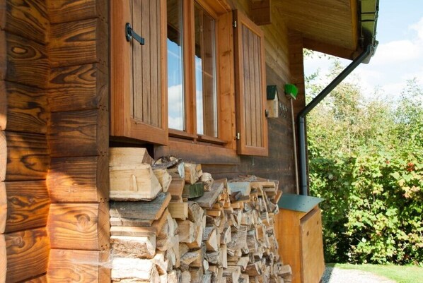 Una cabaña de madera con leña apilada afuera, una ventana con contraventanas y una lámpara verde para exteriores.