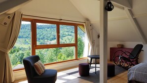 Ein Zimmer mit großem Fenster und Blick auf eine bewaldete Landschaft. Es gibt einen schwarzen Sessel mit Kissen, einen kleinen runden Tisch und eine rote Kommode mit einem Korb darauf.