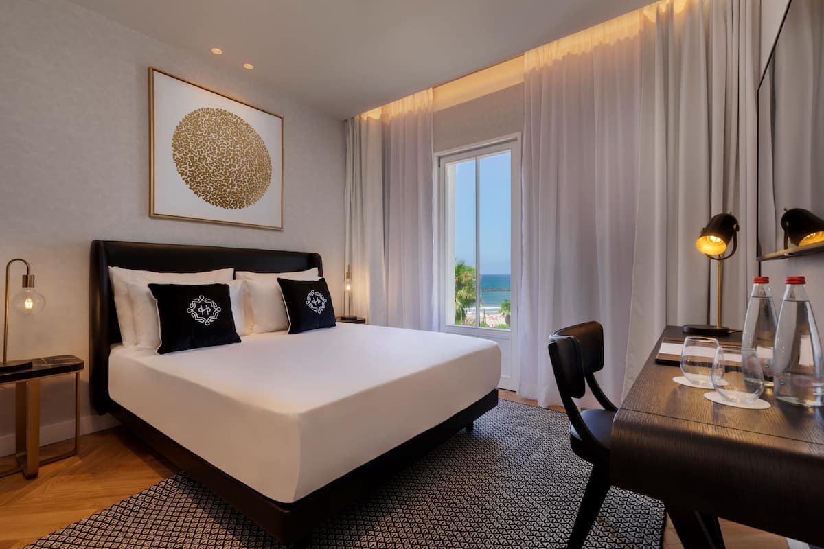 Habitación de hotel con una cama grande, un escritorio con dos vasos y botellas, una mesita de noche con lámpara, y un ventanal con cortinas.
