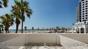 Un balcón con mesa y sillas, con vista a una playa con palmeras y un edificio con la leyenda 