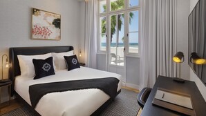 Un cuarto de hotel con una cama grande, un escritorio con lámpara, un cuadro en la pared y vista al mar a través de la ventana.