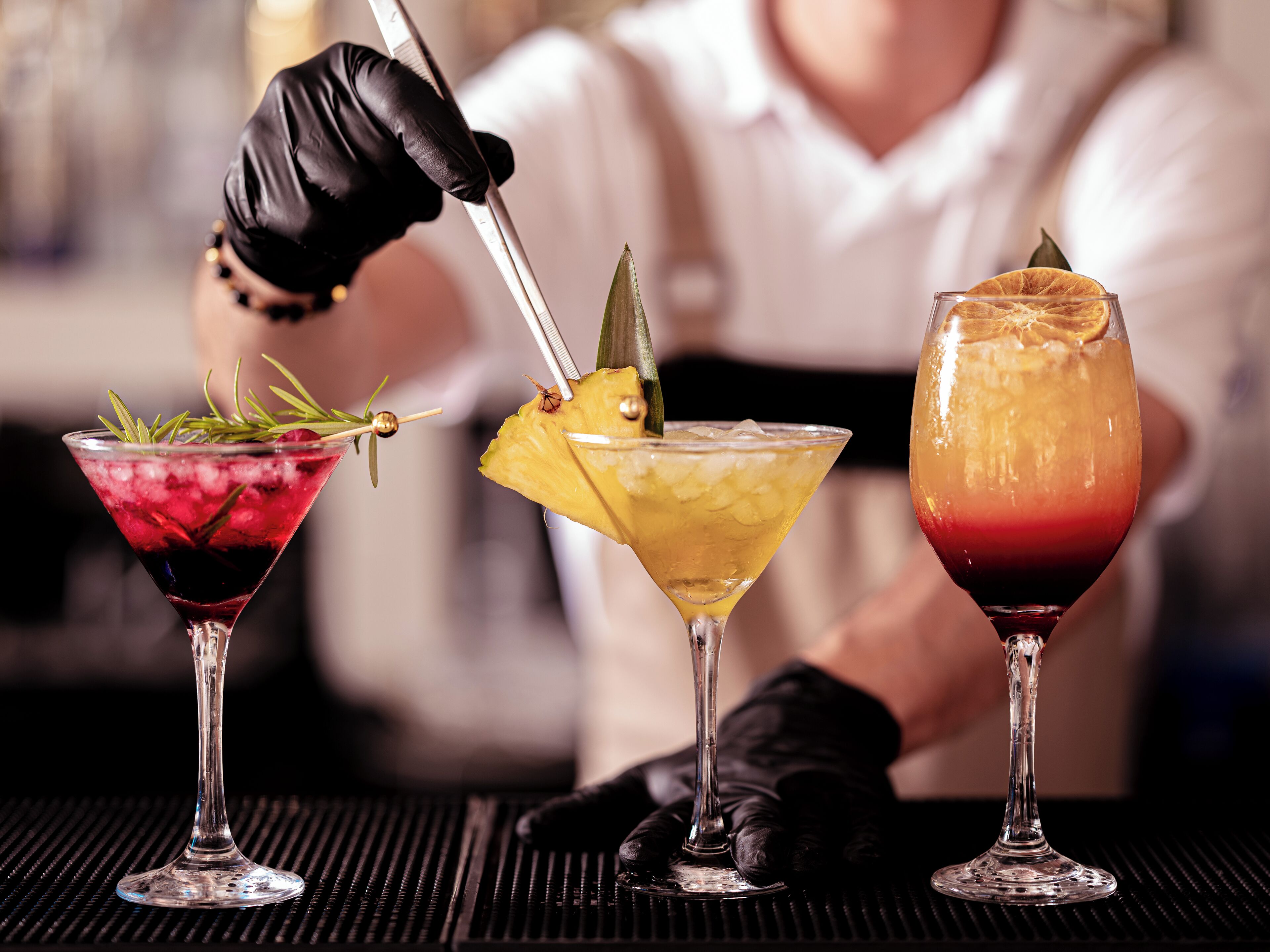 Trois cocktails servis dans des verres à martini, décorés de garnitures, une personne portant des gants noirs qui les prépare, et un arrière-plan flou.