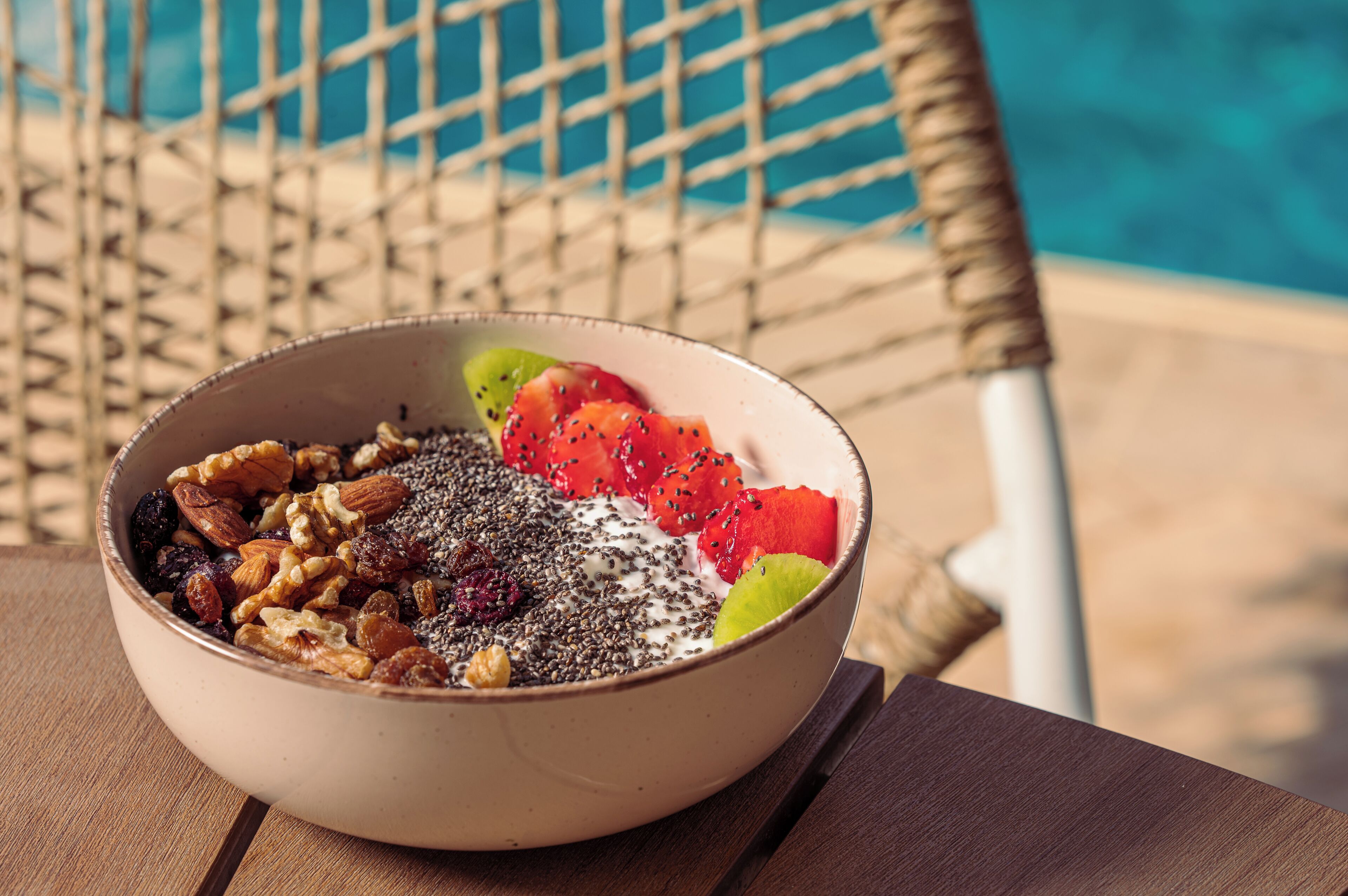 A bowl of mixed nuts and fruits on a wooden table near a poolside chair.