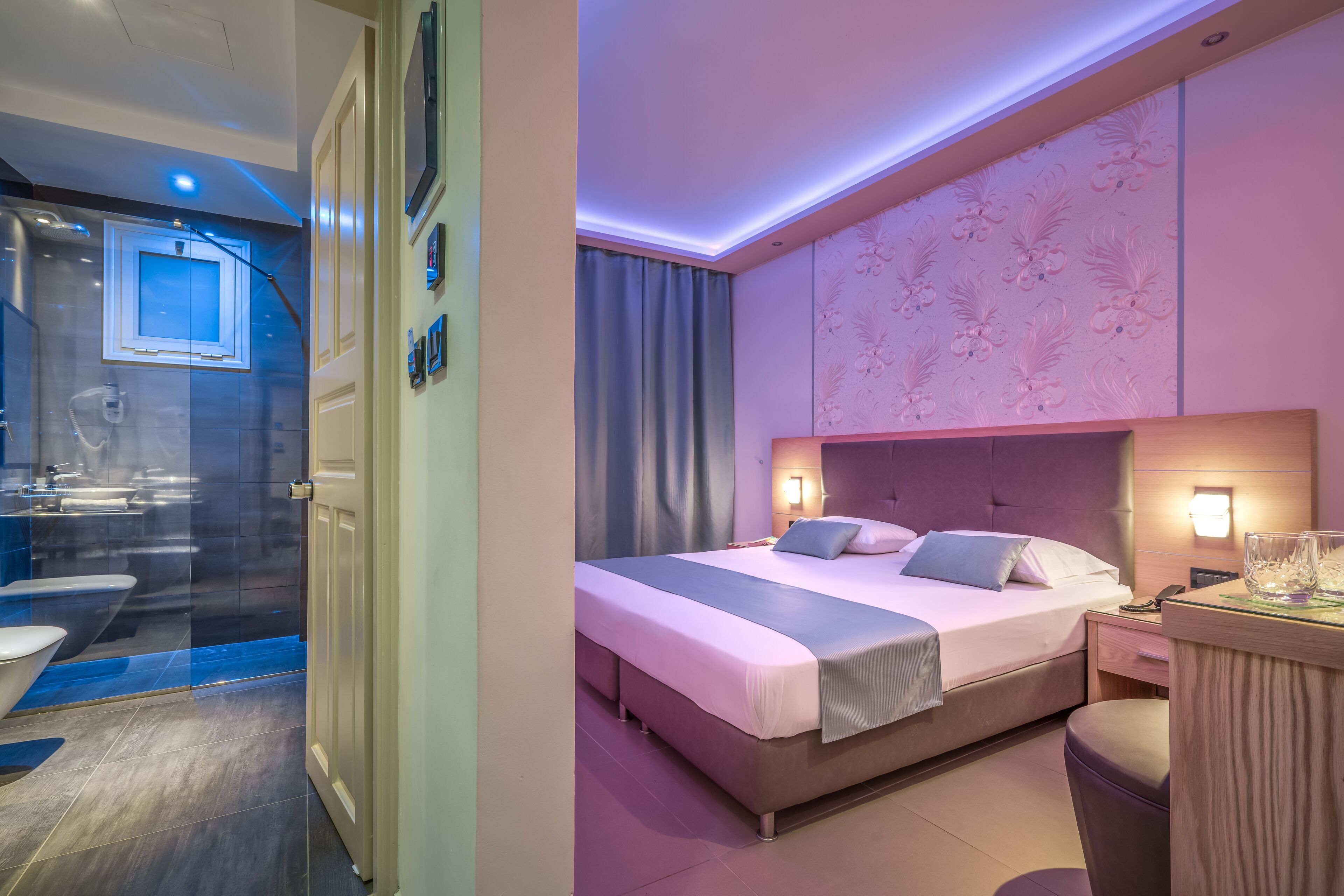 Una camera d'hotel moderna con un letto grande, un comodino con una lampada, un bagno con box doccia in vetro e un pannello decorativo a parete.