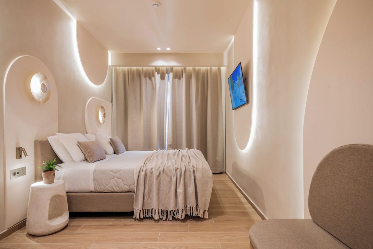 A modern bedroom with a bed, a side table with a plant, a sofa, and a TV on the wall.