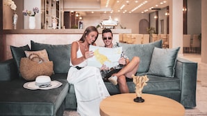 Een koppel zit op een grijze bank in een goed verlichte lobby, met een kaart, een geweven tas en een hoed op de bank.