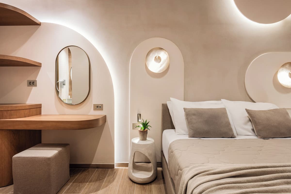 A modern bedroom with a curved vanity, a round mirror, a bedside plant, and a bed with a beige blanket.