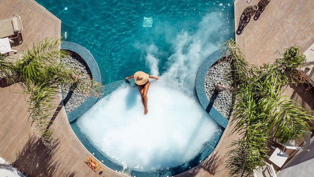 Een rond hottub met een persoon in badpak en hoed, omringd door ligbedden en palmbomen.