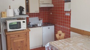 Een compacte keuken met een magnetron, waterkoker en een kleine koelkast. Er is een houten kastje met een klok, een betegelde spatwand en een kalender aan de muur.