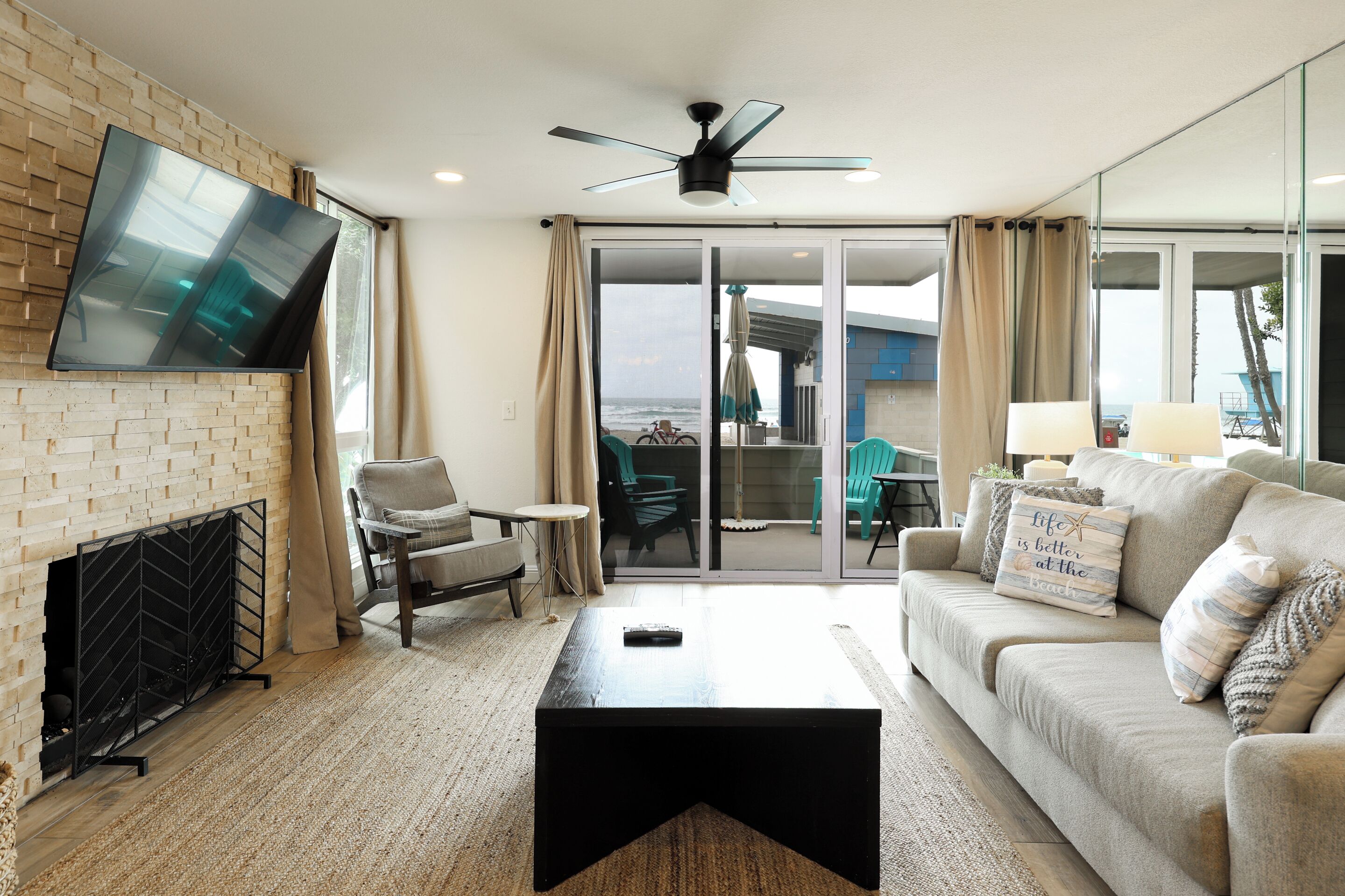 A living room with a fireplace, a television mounted on the wall, a ceiling fan, and a view of the beach through the sliding glass doors.