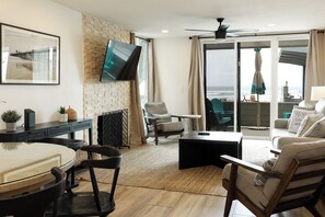 Un salon avec un téléviseur fixé au mur en briques, un coin repas avec une table et des chaises, et un balcon avec vue sur l’océan.