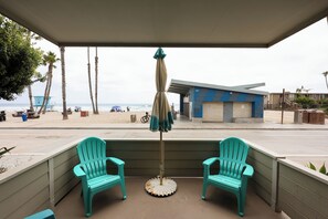 A covered outdoor seating area with two turquoise chairs and a table, overlooking a beach with palm trees and a building with the number 900.