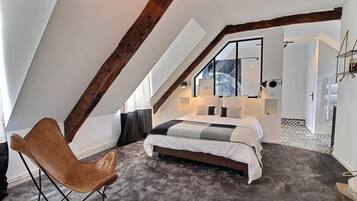 Une chambre avec un plafond en pente, un lit recouvert d’une courtepointe, un fauteuil en cuir et une grande fenêtre.