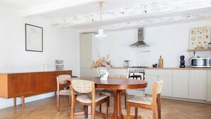 A dining area with a wooden table and chairs, a white cabinet, and a kitchen in the background. - Artefact (Lamballe-Armor)