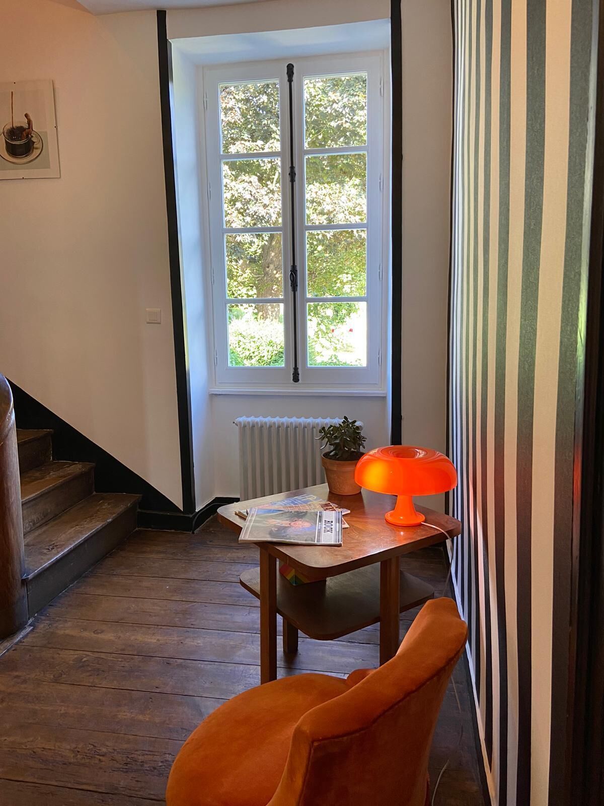 A room with a wooden floor, a window with a view of greenery, a table with a lamp and a plant, and a chair.