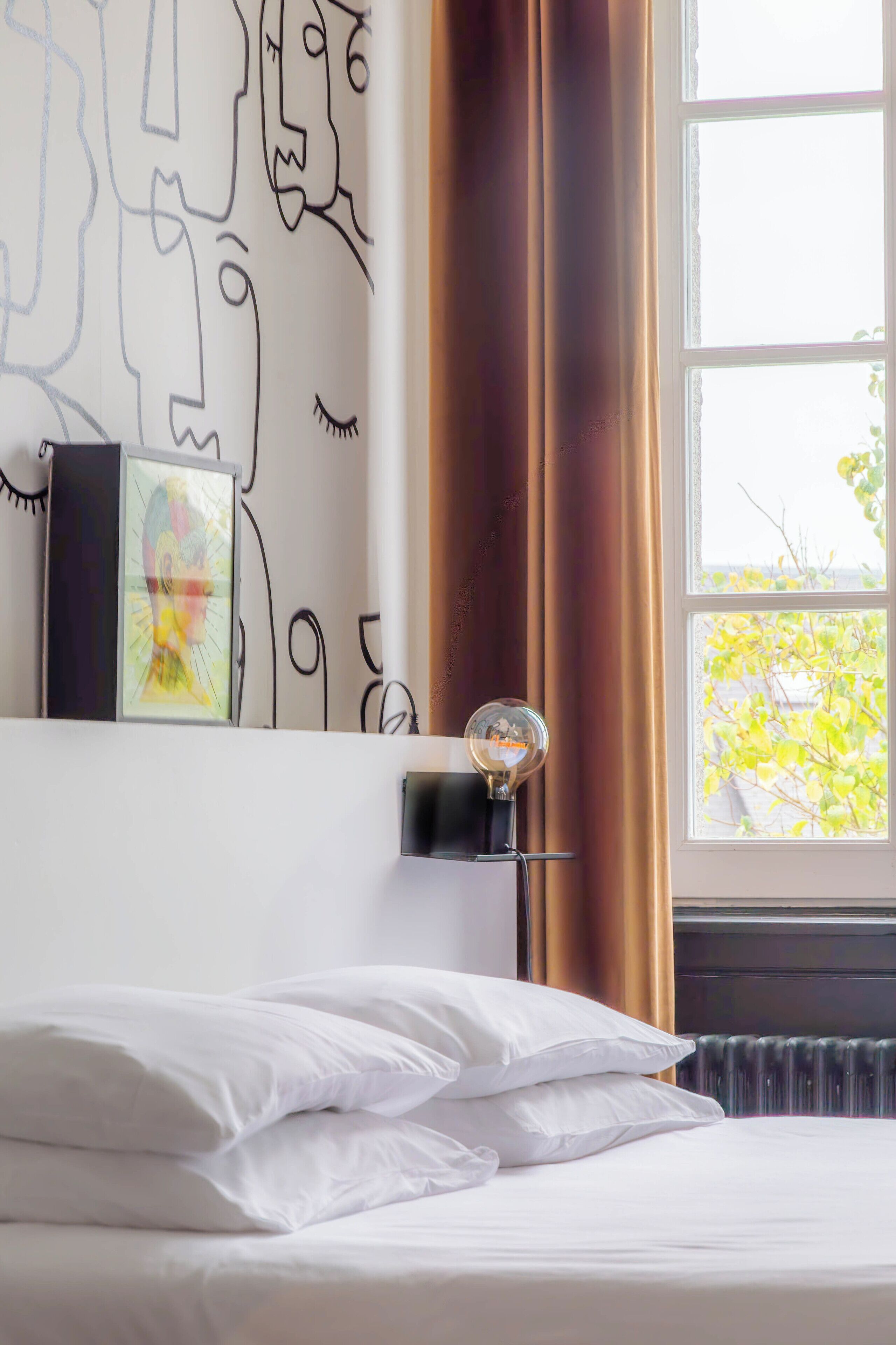 A hotel room with a bed, a wall with abstract art, a window with curtains, and a radiator.