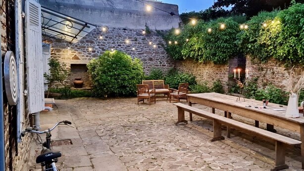 An outdoor patio with stone flooring, wooden furniture, and string lights. - Artefact (Lamballe-Armor)