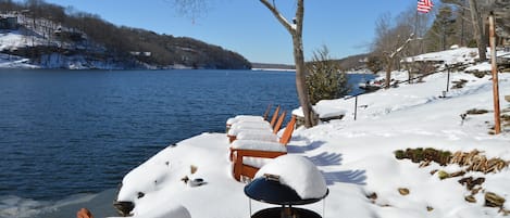 A row of snow-covered Adirondack chairs facing a lake with a clear sky.