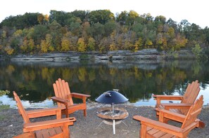 A lakeside setting with two wooden Adirondack chairs and a metal fire pit.