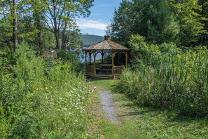 Um gazebo com estrutura de madeira e telhado, rodeado por vegetação exuberante e um caminho de cascalho que o leva até ele.