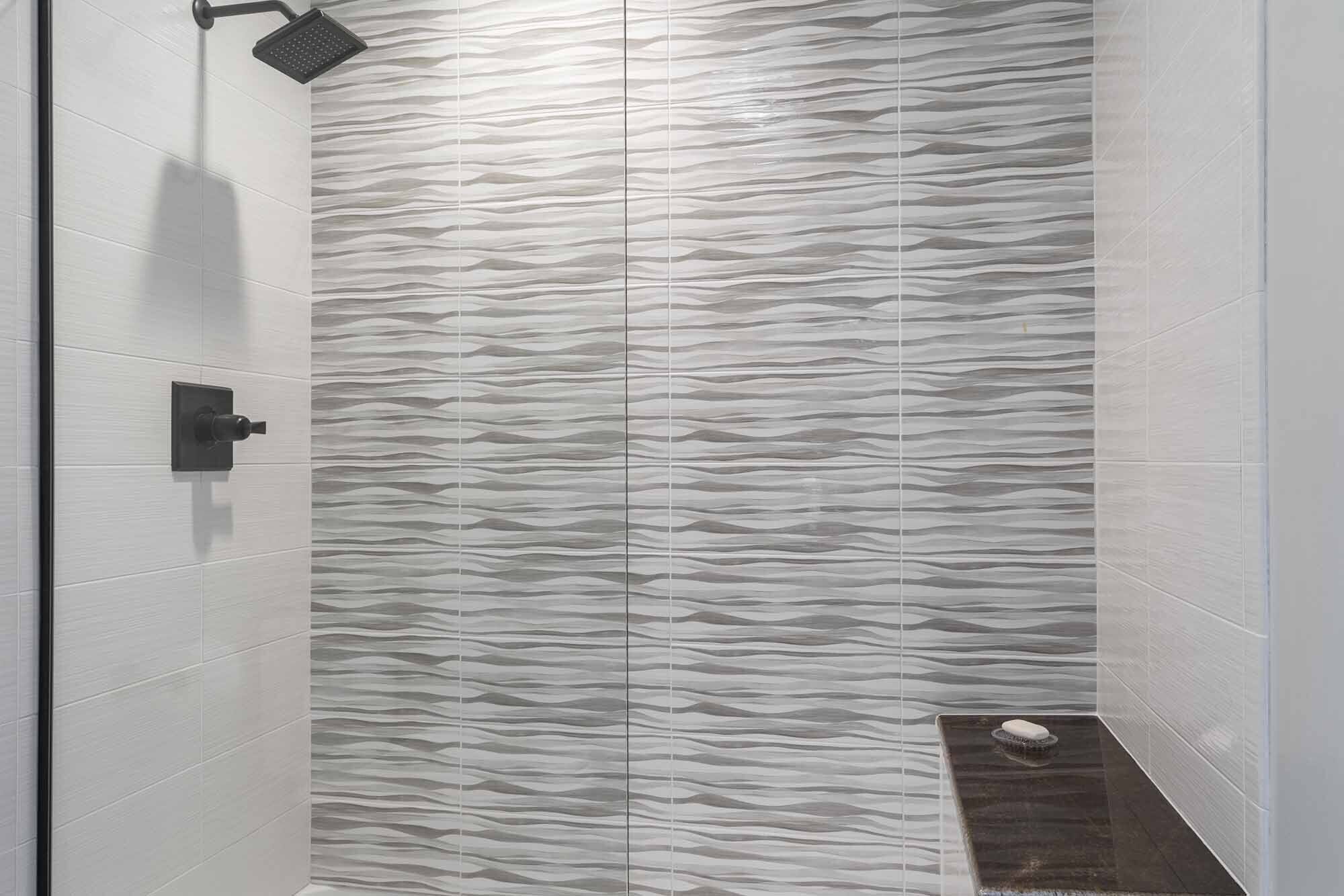Eine Dusche mit wellenförmigem Fliesenspiegel, einer Handbrause und einer kleinen, dunkel gefärbten Fußmatte.