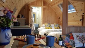 Een gezellig houten vakantiehuisje met een eethoek, keuken en slaapkamer met een bed en kleurrijke kussens.