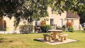 A courtyard with a stone table and benches, surrounded by well-maintained hedges and a building with a tiled roof.