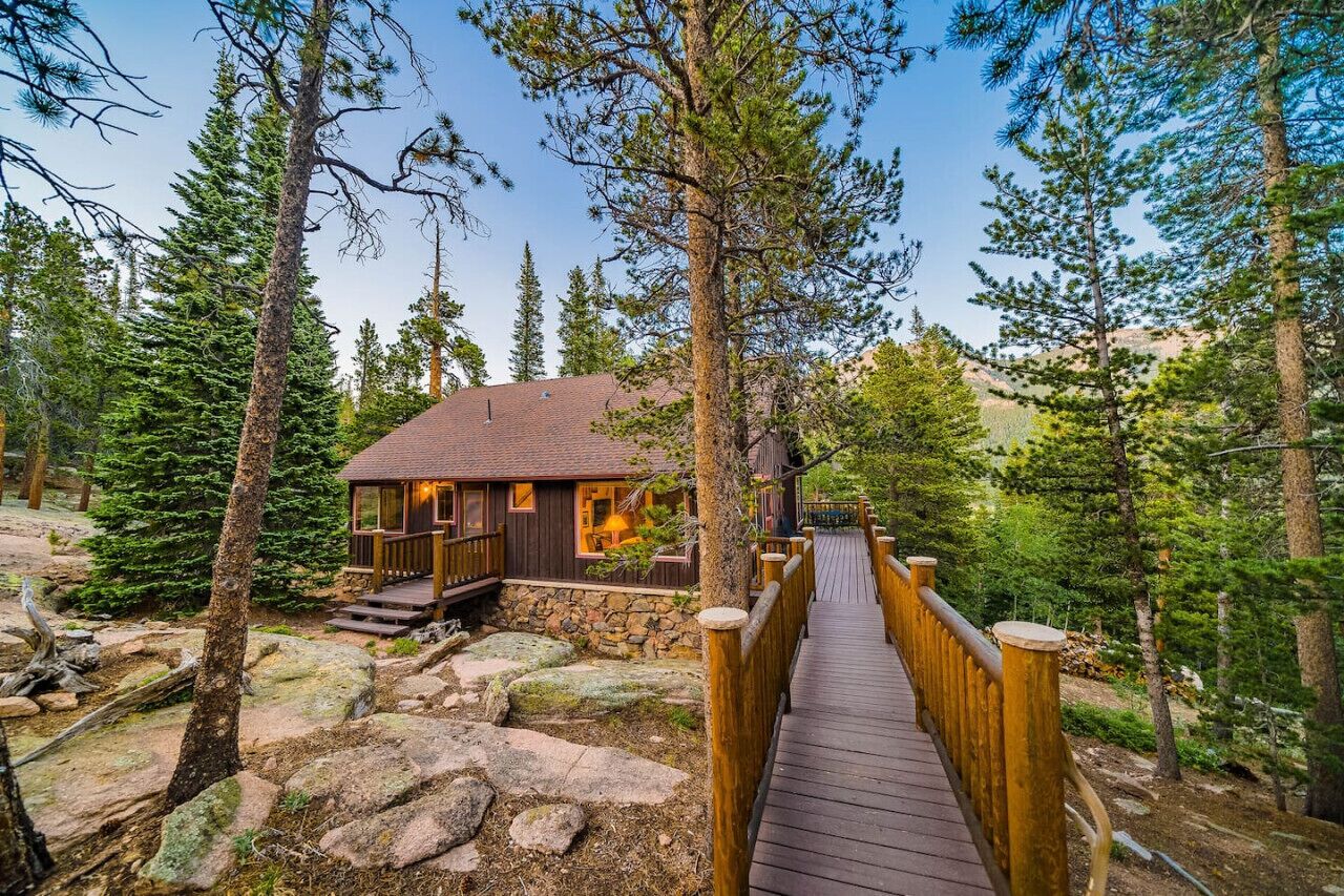 Eine Holzhütte mit einer Holzterrasse, die zu einem Steinfundament führt, umgeben von dichtem Wald.