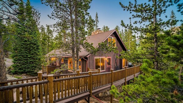 A rustic cabin with a wooden deck and large windows, surrounded by dense forest.