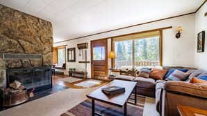 A cozy living room with a stone fireplace, a sofa set, and a large window offering a view of trees.