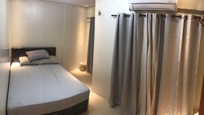 A small, neatly made bed with a headboard, a bedside table with a lamp, and a curtain partially drawn to the side. - Your Home Away From Home (Talisay)