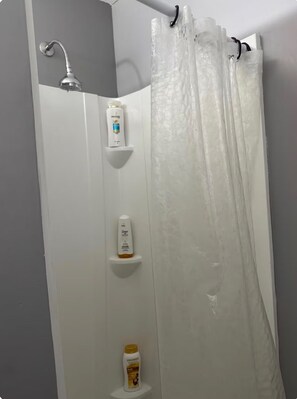 Et bruseområde med brusehoved, et hvidt brusebadeforhæng og tre hylder med toiletartikler.