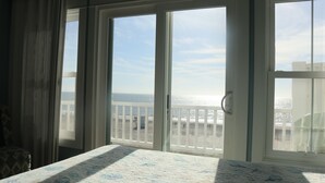 A room with a large window overlooking a beach, a bed with a patterned bedspread, and a view of the ocean. - Ocean Front Ocean View, With Private Heated Pool & Hot Tub (Atlantic Beach)