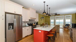 A modern kitchen with a stainless steel refrigerator, a red island with stools, a large window with a view, and a ceiling fan. - Ocean Front Ocean View, With Private Heated Pool & Hot Tub (Atlantic Beach)