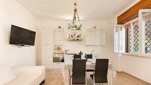 A modern kitchen with white cabinets, a dining table with four chairs, a flat-screen TV mounted on the wall, and a window with a lattice design.