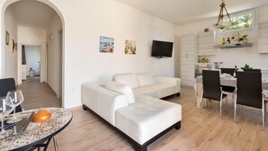 A modern living room with a white sofa, a dining table with chairs, and a kitchen area with a TV and cabinets.