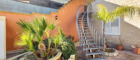 Una escalera de caracol conduce a un segundo piso, rodeada de plantas en macetas y un muro de ladrillo.