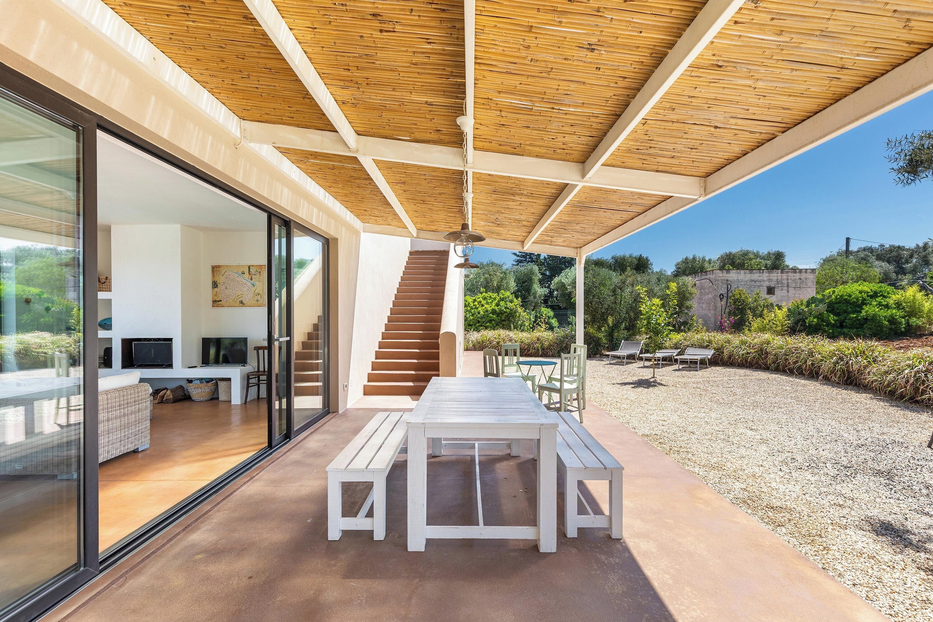 A modern outdoor patio with a wooden ceiling, a white table with benches, and a view of the interior through glass doors.