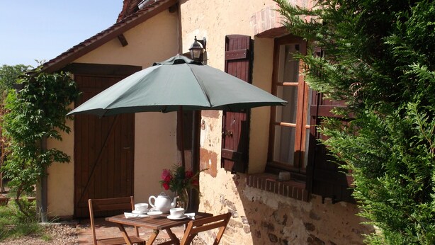 An outdoor wooden table with a green umbrella, surrounded by chairs, set against a rustic stone wall with a window and wooden shutters. - Chez Sidonie, in the land of Colette. (Treigny-Perreuse-Sainte-Colombe)