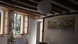 A room with a wooden ceiling, a white wall with a landscape painting, a sofa, a table, and a window with open shutters. - Chez Sidonie, in the land of Colette. (Treigny-Perreuse-Sainte-Colombe)